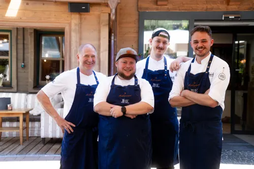 Four smiling chefs in blue aprons standing outside a rustic restaurant